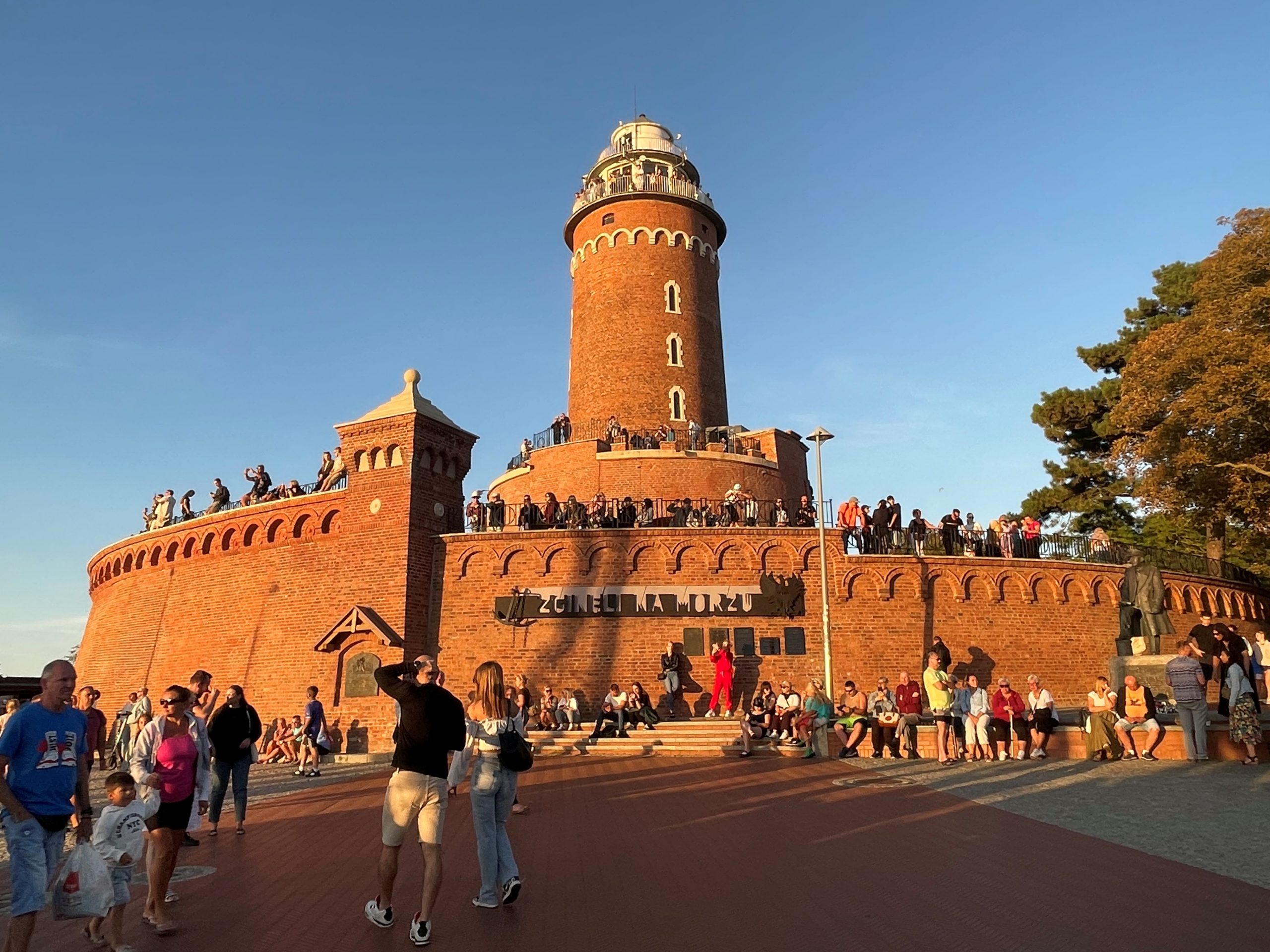 latarnia morska w Kołobrzegu, ludzie spacerują, chodzą wokół zabytku, siedzą